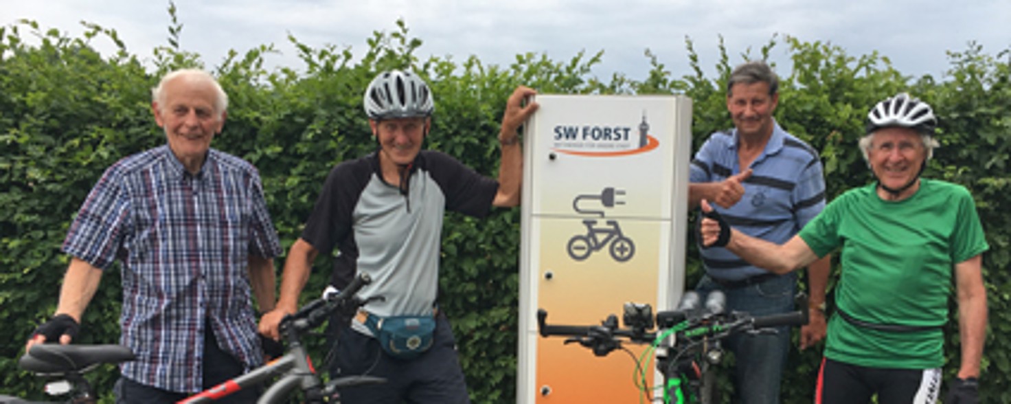 Vier ältere Herren in Fahrrad-Outfits stehen mit ihren Fahrrädern neben einer Fahrrad-Ladestation