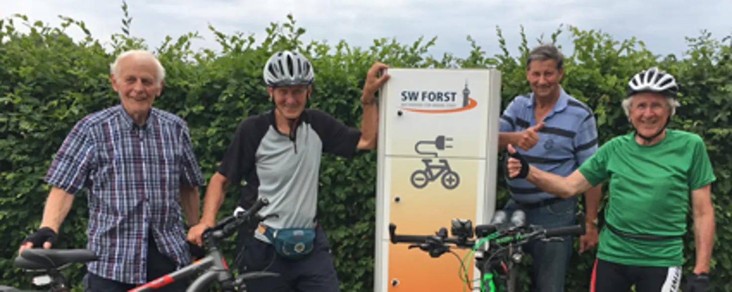 Vier ältere Herren in Fahrrad-Outfits stehen mit ihren Fahrrädern neben einer Fahrrad-Ladestation