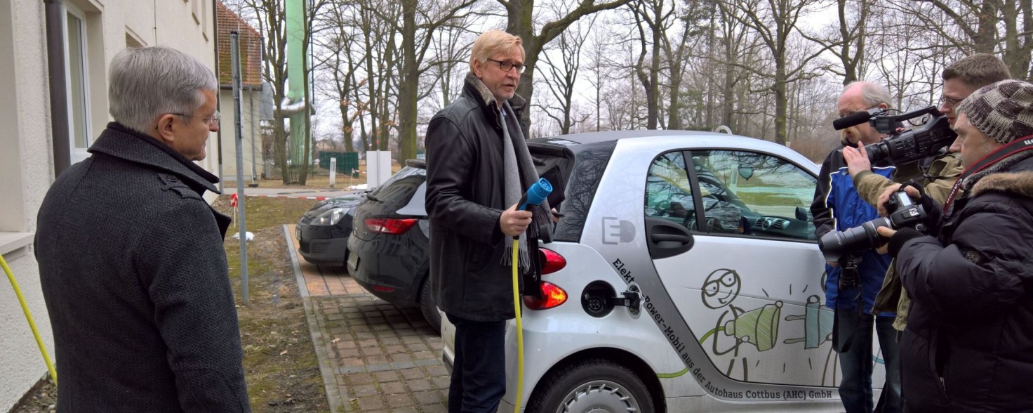 Ein Mann steht mit einem Ladekabel neben einem E-Auto und wird von einem Kamerateam gefiltm