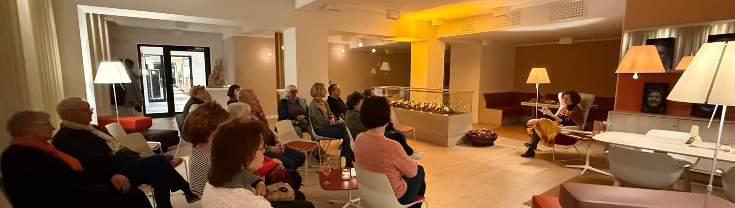 Menschen sitzen auf Stühlen vor einer Frau im Sessel die vorliest