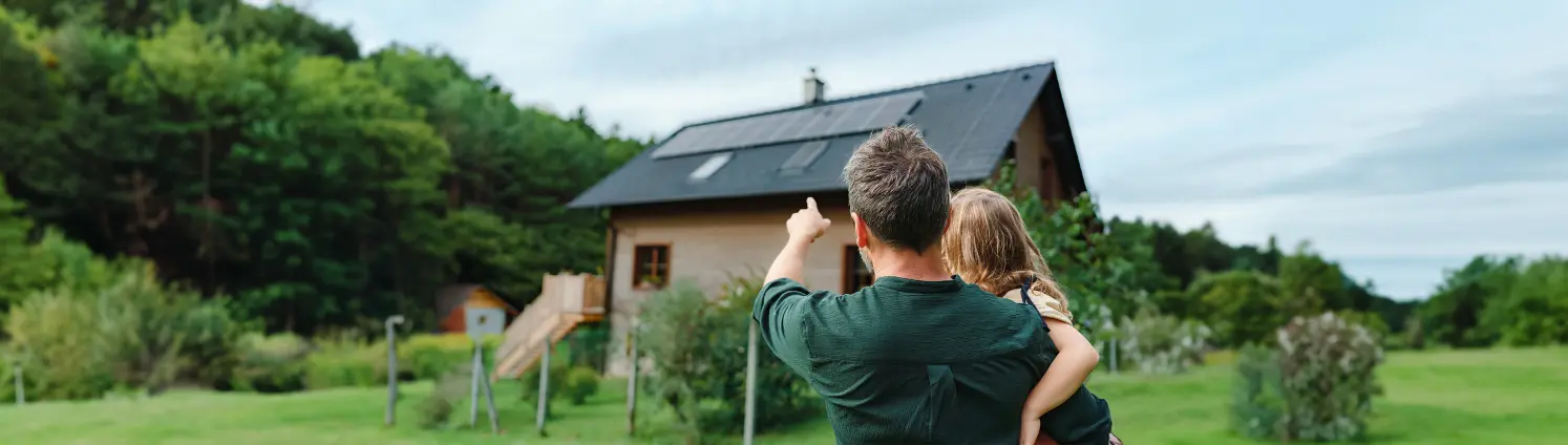 Rückansicht eines Mannes, der seine Tochter auf dem Arm hält und auf ein Haus mit Sonnenkollektoren zeigt. Bild mit KI erstellt.