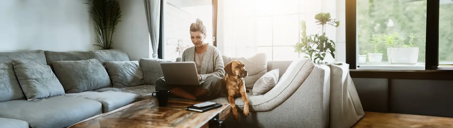 Geschäftsfrau sitzt mit ihrem Hund auf der Couch und arbeitet am Laptop. Bild mit KI erstellt.