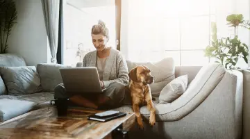 Geschäftsfrau sitzt mit ihrem Hund auf der Couch und arbeitet am Laptop. Bild mit KI erstellt.